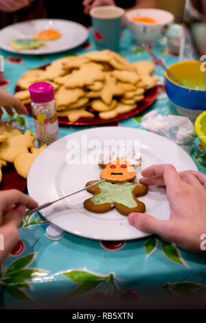 Nahaufnahme, Hände ein Kids in den Prozess der Verzierung eines frisch gebackenen Plätzchen wie ein Rentier mit grün und orange Sahnehäubchen auf eine weisse Platte geformt Stockfoto