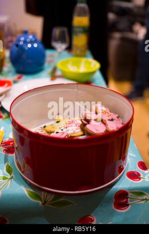 Eine Keksdose voll mit frisch gebackenen und hausgemachte dekoriert Weihnachten Kekse mit Zuckerguss, Konfetti und Glitter bedeckt sitzt auf einem bunten Tisch bei Kaffee Stockfoto
