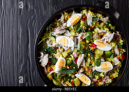 Kedgeree mit gehobelten Fische, geräuchert, hart gekochte Eier, Reis, Kohl, Rosenkohl, Gewürze und Kräuter in einem keramischen Pan? Auf einer hölzernen Tabelle, Ansicht von oben Stockfoto