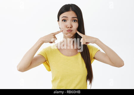 Portrait von Silly feminin und weiblich Modell in gelben T-Shirt schmollen, Atem und stossen Wangen mit Index Finger, lächerlich und lustig Spaß und Aprilscherze über graue Wand Stockfoto