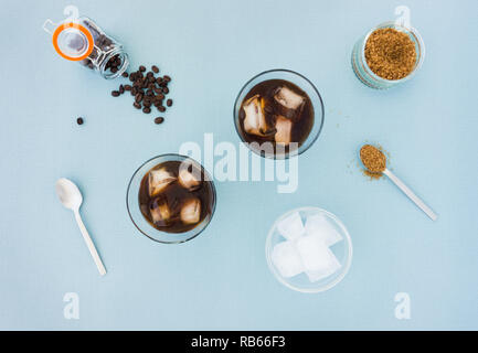 Gläser Eiskaffee, Kaffee Bohnen, Zucker und eine Schale mit Eiswürfeln auf blauem Hintergrund gefüllt. Ansicht von oben. Stockfoto