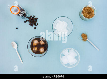 Gläser kalter Kaffee brühen, Kaffeebohnen, Zucker und eine Schale mit Eiswürfeln auf blauem Hintergrund gefüllt. Ansicht von oben. Stockfoto