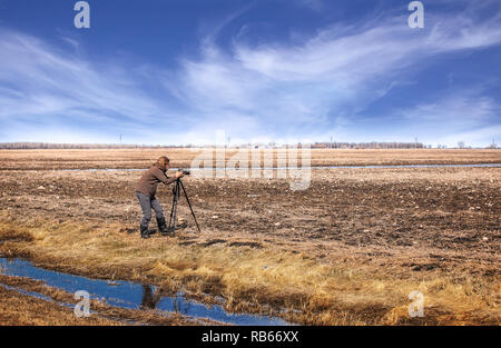 Eine Frau in dünne Jacke und Gummistiefel einrichten Stativ und Kamera Fotos in einer braunen nasser Frühling Landwirtschaft Feld zu nehmen Stockfoto