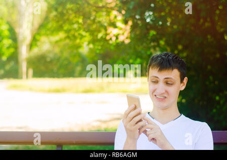 Eine junge europäische Kerl sitzt auf einer Bank im Stadtpark und macht ein lustiges Gesicht schauen in das Telefon. Das Konzept der extremen überraschen und Ekel, plötzliche Stockfoto