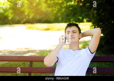 Eine junge europäische Kerl sitzt auf einer Bank im Park und am Telefon zu sprechen, wirft seinen Arm hinter den Kopf und schließen die Augen. Entspannt Telefon Stockfoto