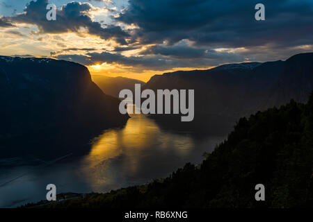 Einen goldenen Sonnenuntergang über Felswände in den Fjord Gewässern, unter einem bewölkten Himmel. Norwegen, Aurlandsfjord. Stockfoto