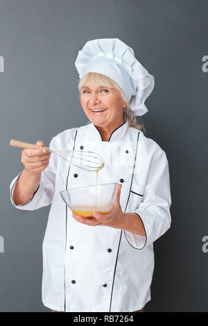 Senior Köchin Studio stehen isoliert auf graue Wand holding Schneebesen mischen Eier in der Schale, die Kamera lächelt Fröhlich Stockfoto