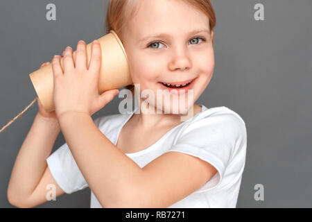 Kleine Mädchen stehen isoliert auf graue Wand spielen mit blechdose Telefon lächeln schließen aufgeregt - Bis Stockfoto