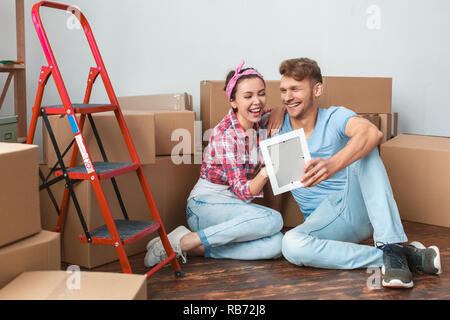 Junge Mann und Frau, der seinen neuen Platz sitzen auf dem Boden und umarmte Holding Bilderrahmen lachen Fröhlich lustiger Moment Stockfoto