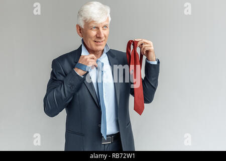 Senior Business mann Studio stehen isoliert auf graue Wand Wahl binden Farbe versuchen, auf beiseite schaut lächelnd nachdenklich Stockfoto