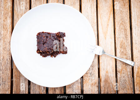 Flach auf eine vegane Chocolate Brownie Festlegung auf einer weißen Platte auf einem Holztisch und eine Gabel Stockfoto