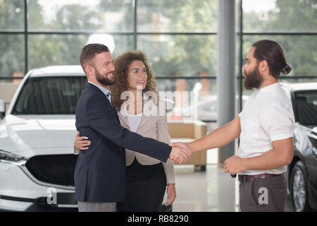 Paare, die sich mit Manager Autohaus Automobil kaufen. Bärtiger Mann Händeschütteln mit Manager und an jedem anderen suchen. Schöne lächelnde Frau mit der Hand runde Mann Taille. Stockfoto