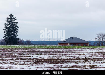 Ein bisschen Schnee auf einem gepflügten Feld und Weide. Haus und im Hintergrund Fichte. Beginn des Winters in Europa. Stockfoto