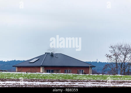 Ein bisschen Schnee auf einem gepflügten Feld und Weide. Haus, Baum und Wald im Hintergrund. Beginn des Winters in Europa. Stockfoto