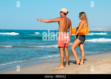 Weibliche und männliche Rettungsschwimmer mit Ausrüstung am Strand gegen den klaren Himmel. Männliche Rettungsschwimmer zeigt mit dem Finger in die Ferne. Kreta. Griechenland. Stockfoto