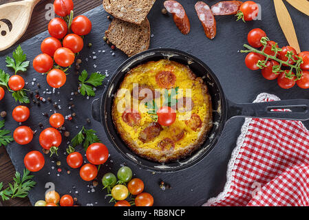 Spiegeleier omelette von Huhn Eier mit roten Kirschtomaten und geräucherte Wurst in einer Runde, schwarze Pfanne, Ansicht von oben Stockfoto