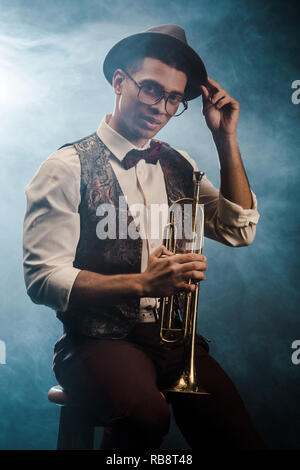 Schöne stilvolle junger Mann in Hut und Brille mit Trompete auf der Bühne mit Rauch und dramatische Beleuchtung posing Stockfoto