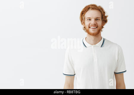 Taille-up Shot sorglosen gewöhnlichen redhead bärtiger Mann mit unordentlichen stilvolle Frisur in weiss Poloshirt lächelnd mit Casual freundlich und glücklich lächeln genießen großartige Sommertag über grauer Hintergrund Stockfoto