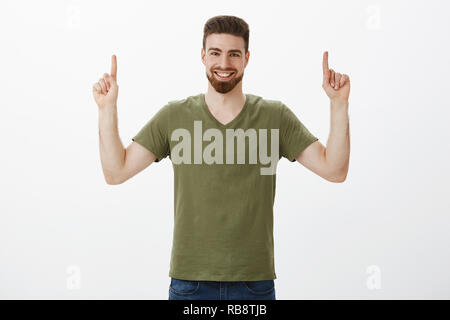 Taille-up Shot der Charismatischen kaukasischen Mann mit Bart in Oliv t-shirt heben die Hände oben mit Index Finger. lächelt zufrieden und begeistert von der Kamera mit glücklichen Ausdruck über weiße Wand Stockfoto