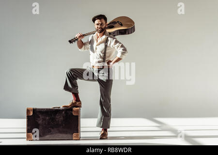 Bärtiger Mann im Cap mit Vintage Koffer mit akustischer Gitarre und Kamera auf grauem Hintergrund Stockfoto