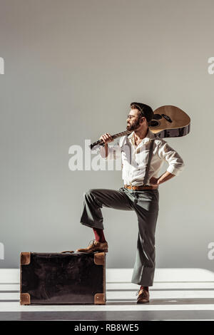 Bärtiger Mann in Gap, Fuß auf vintage Koffer und mit akustischer Gitarre über der Schulter auf grauem Hintergrund Stockfoto