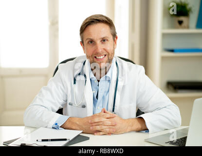 Portrait von Happy kaukasischen Arzt im weißen Kittel Arbeiten am Laptop und Unterlagen in Klinik Krankenhaus Schreibtisch für Health Care erfolgreiche professio Stockfoto