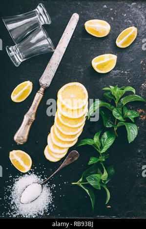 Blick von oben auf die Cocktail Zutaten Zubereitung auf schwarzem Hintergrund im Landhausstil. Tequila Shot Gläser, Zitronenscheibe, Salz und Minze auf grunge dunklen Tabelle Form ab Stockfoto