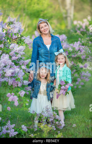 Mutter Frau mit zwei nett lächelnden Mädchen Schwestern gemeinsam schön auf einem lila Feld Bush alle tragen elegante Kleider und Mäntel Jeans Stockfoto