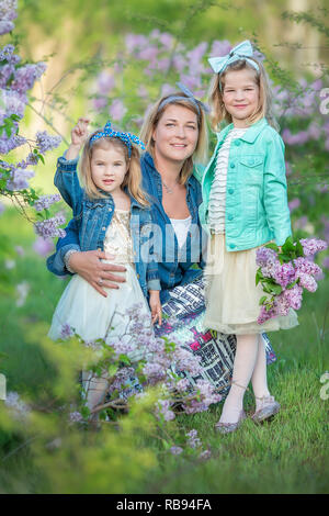 Mutter Frau mit zwei nett lächelnden Mädchen Schwestern gemeinsam schön auf einem lila Feld Bush alle tragen elegante Kleider und Mäntel Jeans Stockfoto