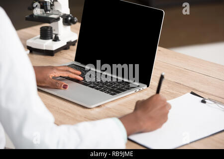 7/8-Ansicht der afrikanischen amerikanischen Arzt Schreiben auf Papier und mit Laptop mit leerer Bildschirm Stockfoto