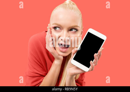 Porträt einer überrascht, lächelnd, glücklich, erstaunt Mädchen mit leerer Bildschirm Mobiltelefon über Coral Hintergrund isoliert. Stockfoto