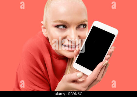 Porträt einer überrascht, lächelnd, glücklich, erstaunt Mädchen mit leerer Bildschirm Mobiltelefon über Coral Hintergrund isoliert. Stockfoto