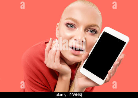 Porträt einer überrascht, lächelnd, glücklich, erstaunt Mädchen mit leerer Bildschirm Mobiltelefon über Coral Hintergrund isoliert. Stockfoto