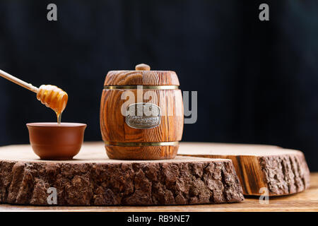 Geschlossene Holz Fass mit Honig und Honig Löffel auf einer hölzernen Säge auf einem dunklen Hintergrund. Barrel. Kopieren Sie Platz. Close-up. Stockfoto