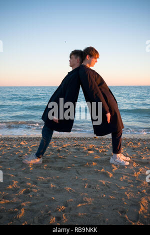 Zwei Jungen im Teenageralter tragen ähnliche Kleidung Narr über am Strand von gegeneinander gelehnt wie die Sonnenuntergänge im südlichen Frankreich Stockfoto