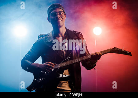 Lachende junge männliche Musiker in Lederjacke auf der E-Gitarre auf der Bühne mit Rauch und dramatische Beleuchtung Stockfoto