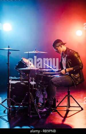 Männliche Rockstar in Leder Jacke Schlagzeug spielen beim Konzert auf der Bühne mit Rauch- und Strahler Stockfoto