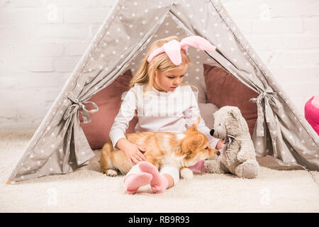 Kind in hasenohren kopfband Sitzen mit corgi Hund und Teddybären im Wigwam zu Hause Stockfoto