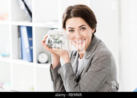 Attraktive happy Geschäftsfrau holding Glas Glas mit Geld im Büro und suchen an Kamera Stockfoto