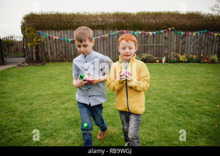 Zwei Jungen zu Fuß durch den Garten nach einem Easter Egg Hunt. Sie halten eine Handvoll Schokolade Eier in ihren Händen. Stockfoto