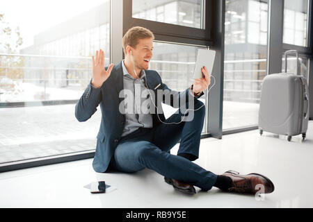 Fröhlicher junger Mann auf dem Boden sitzen und Gruß. Er Wellen mit ahnd. Junge Mann hat einen Videoanruf. Er verwendet die Kopfhörer. Guy blatt Koffer und Telefon mit tic Stockfoto