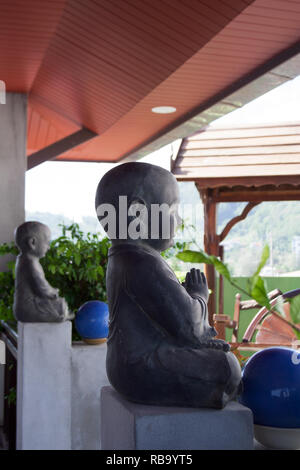 Zwei beten Buddha Skulpturen auf Thai terrasse Zaun mit Pflanzen, blauen Kugeln und hölzerne Schwingen auf dem Hintergrund Stockfoto