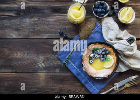 Frische hausgemachte Niederländische Baby Pfannkuchen mit Zitronen Quark und Blaubeeren in Iron Skillet auf rustikalen Holzmöbeln Hintergrund. Ansicht von oben. Kopieren Sie Platz. Stockfoto