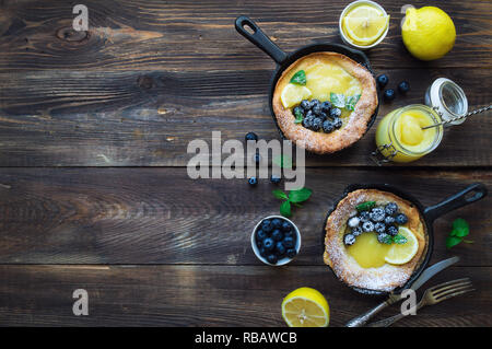 Frische hausgemachte Niederländische Baby Pfannkuchen mit Zitronen Quark und Blaubeeren in Eisen skillets auf rustikalen Holzmöbeln Hintergrund. Ansicht von oben. Kopieren Sie Platz. Stockfoto