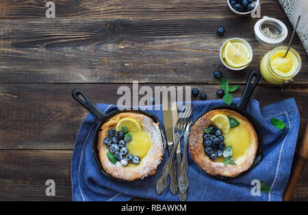 Frische hausgemachte Niederländische Baby Pfannkuchen mit Zitronen Quark und Blaubeeren in Eisen skillets auf rustikalen Holzmöbeln Hintergrund. Ansicht von oben. Kopieren Sie Platz. Stockfoto