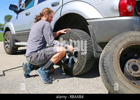 Einem jungen, langhaarigen Mann ist mit Gewalt zu ändern, ist Reifenpanne auf seinem silbernen SUV, mit seiner Freizeit zu sitzen, auf der Seite der Straße geblasen. Stockfoto
