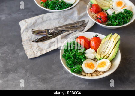 Quinoa buddha Schüsseln mit gebackenen Tomaten, Avocado, Grünkohl, gekochte Eier und griechischer Joghurt auf grauem Beton Hintergrund. Gesundes Frühstück. Stockfoto