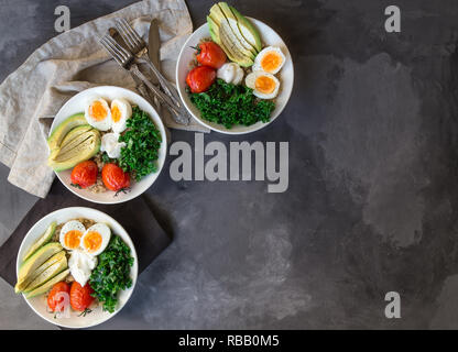 Quinoa buddha Schüsseln mit gebackenen Tomaten, Avocado, Grünkohl, gekochte Eier und griechischer Joghurt auf grauem Beton Hintergrund. Gesundes Frühstück. Ansicht von oben. Kopieren Sie sp Stockfoto