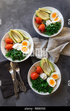 Quinoa buddha Schüsseln mit gebackenen Tomaten, Avocado, Grünkohl, gekochte Eier und griechischer Joghurt auf grauem Beton Hintergrund. Gesundes Frühstück. Ansicht von oben. Stockfoto