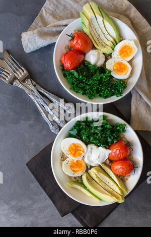 Quinoa buddha Schüsseln mit gebackenen Tomaten, Avocado, Grünkohl, gekochte Eier und griechischer Joghurt auf grauem Beton Hintergrund. Gesundes Frühstück. Ansicht von oben. Stockfoto